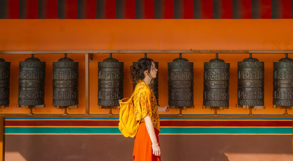 A woman in a yellow shirt and orange skirt walks past a row of prayer wheels against a vibrant orange and turquoise wall.