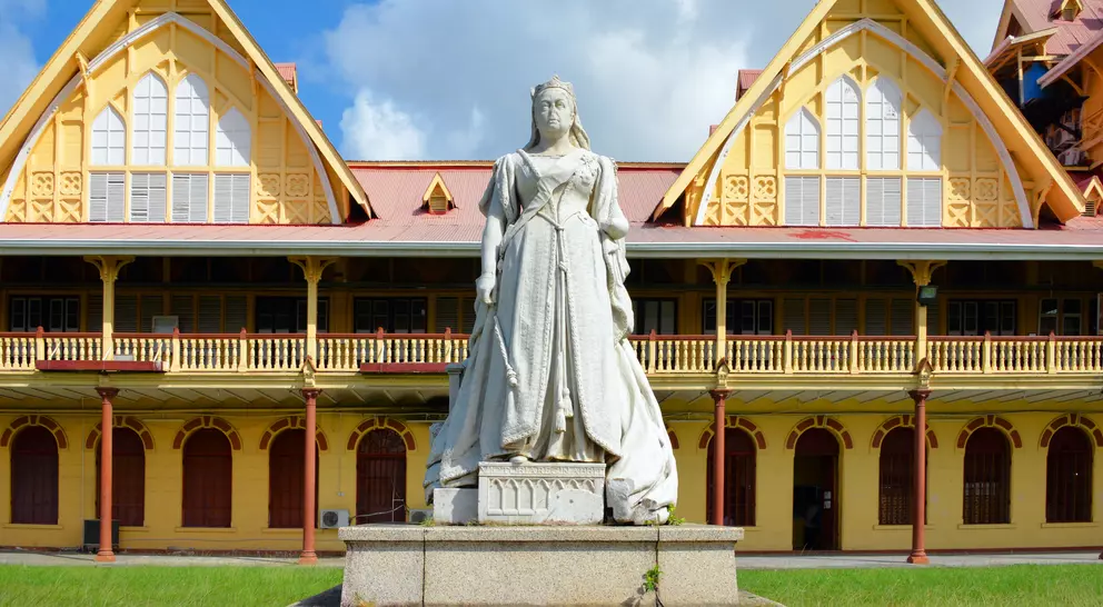 Statue of a woman in white dress in front of a historic yellow building with peaked roofs and blue sky.