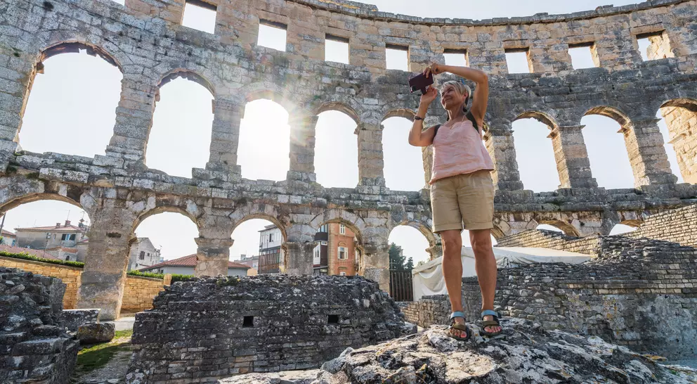 A person stands on rocks, photographing the ruins of a coliseum with sunlight streaming through the arches.
