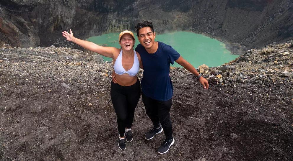 Two smiling people pose near a volcanic crater, with turquoise water visible below, against a dramatic rocky landscape.