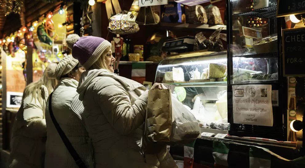 Two women in winter coats browse a festive market stall displaying various cheeses, surrounded by holiday lights.