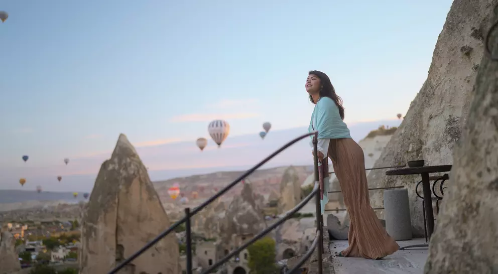 A woman stands on a balcony overlooking a valley with hot air balloons in the sky at sunset.