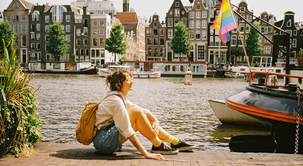 Person sitting by a canal in Amsterdam with colorful buildings and a rainbow flag in the background on a sunny day.