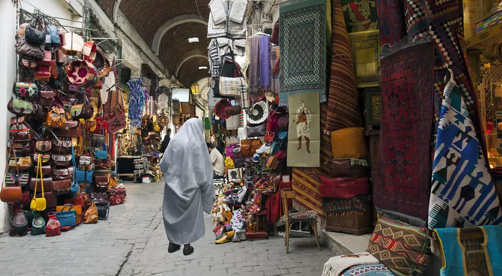 A bustling market with vibrant textiles, bags, and crafts, featuring a person in a gray cloak walking through the narrow path.
