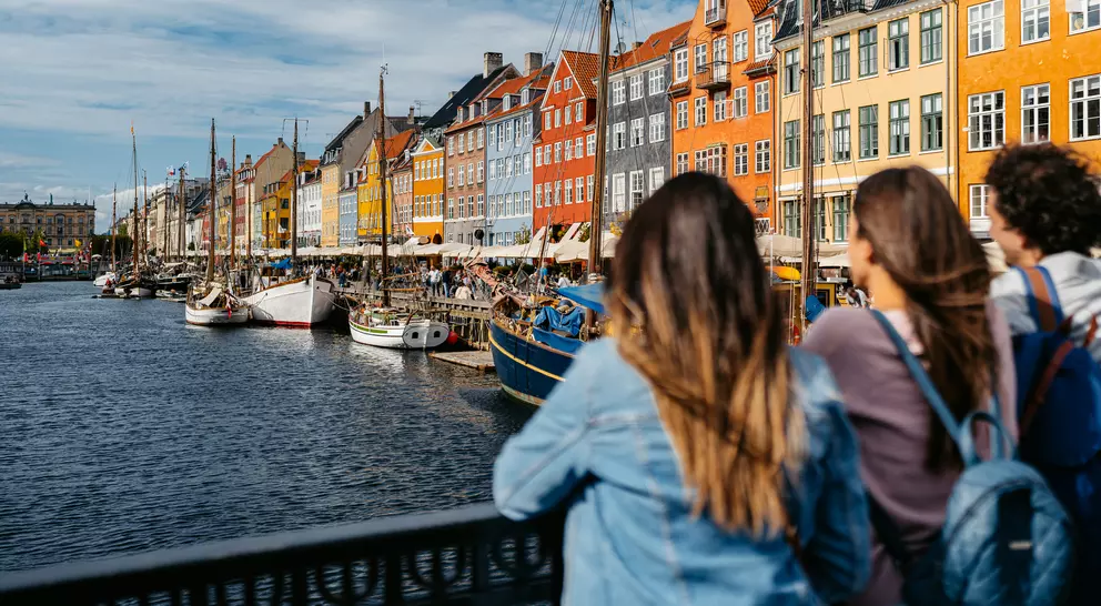 Three people stand by a canal, gazing at colorful buildings and boats in a scenic waterfront view.