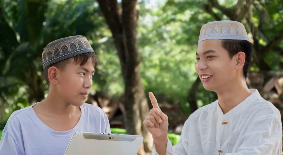 Two young men wearing traditional hats engage in conversation outdoors, one holding a tablet and listening intently.
