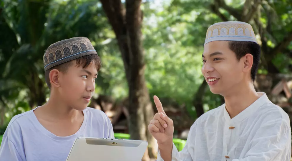 Two young men wearing traditional hats engage in conversation outdoors, one holding a tablet and listening intently.