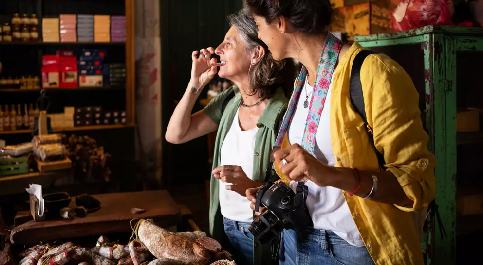 Two women enjoy a moment in a market, surrounded by various cured meats and goods, one holding a camera.