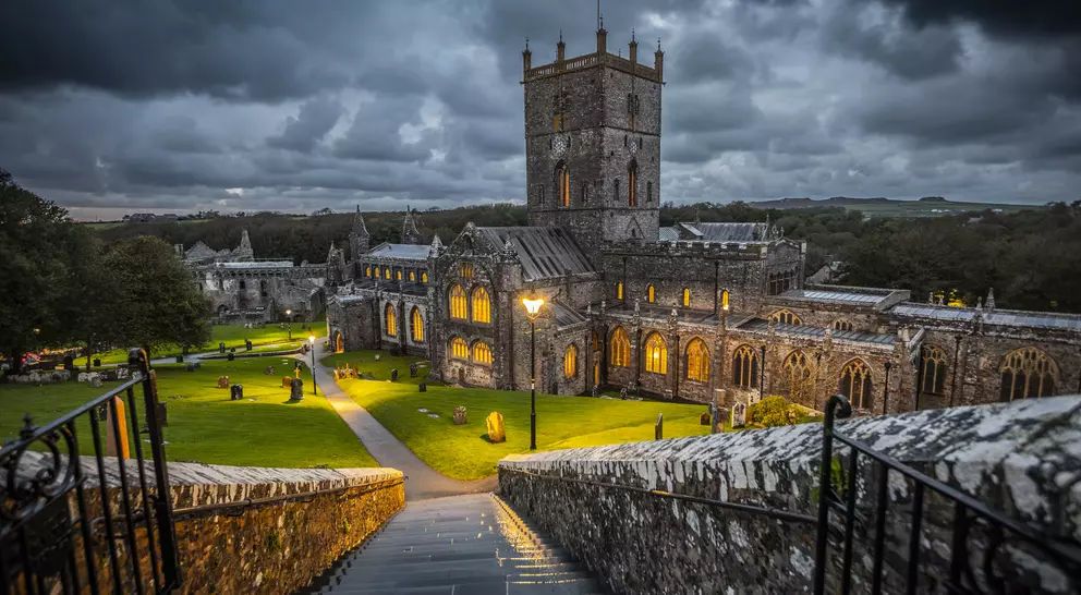 Historic stone church with a tall tower, surrounded by trees and glowing lights, under a dramatic cloudy sky.