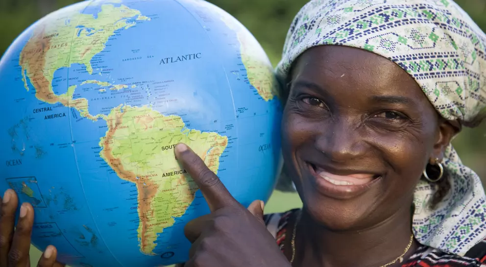Smiling woman pointing at a globe, highlighting South America, against a natural backdrop.