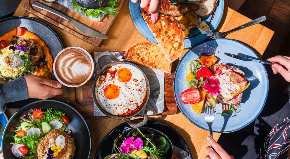 A top view of a diverse brunch spread with dishes, drinks, and hands sharing food around a wooden table.