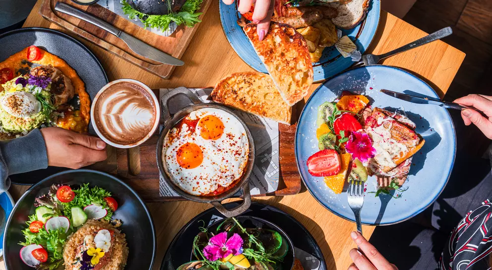 A top view of a diverse brunch spread with dishes, drinks, and hands sharing food around a wooden table.
