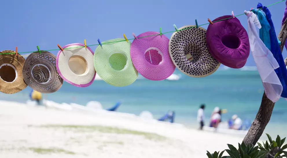 Colorful hats hang on a line with a beach and ocean in the background, accompanied by people enjoying the scenery.