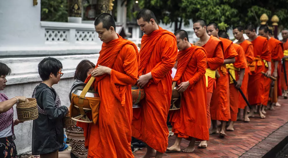 Buddhist monks in orange robes receive alms from followers in a serene outdoor setting.