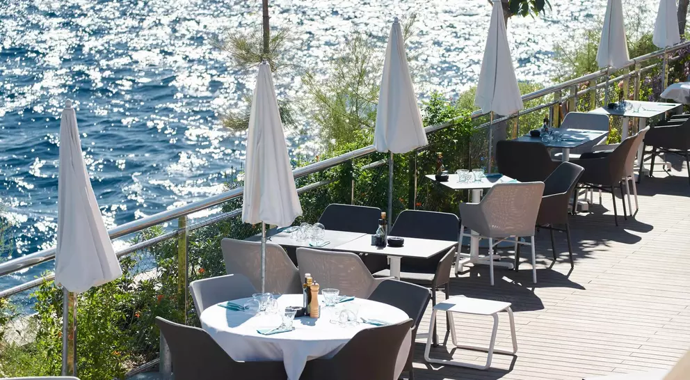 Outdoor restaurant patio with tables and umbrellas beside a sparkling blue sea.