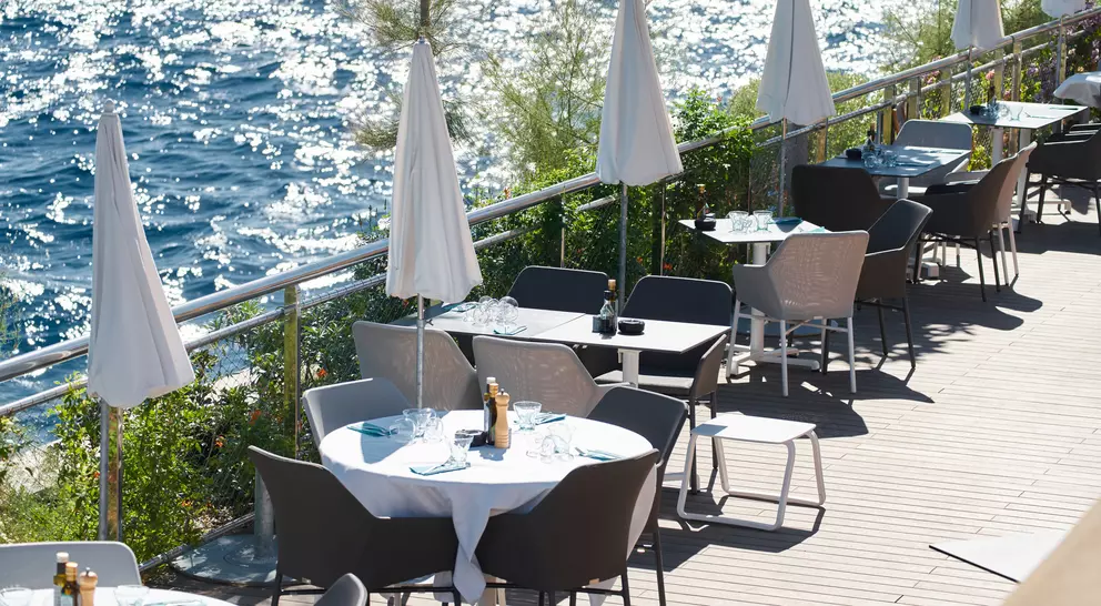 Outdoor restaurant patio with tables and umbrellas beside a sparkling blue sea.