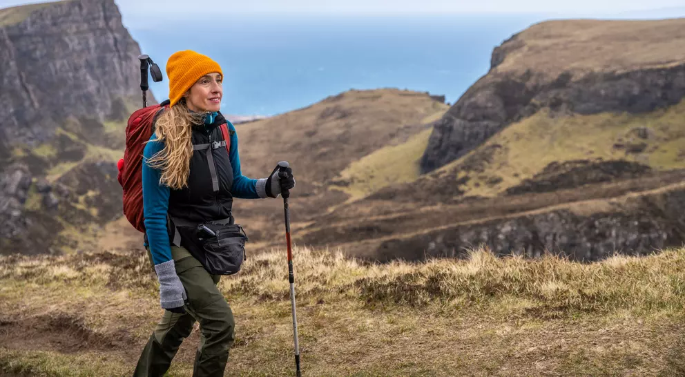 A woman with long hair in a bright orange hat hikes along a coastal path, with cliffs and the ocean in the background.