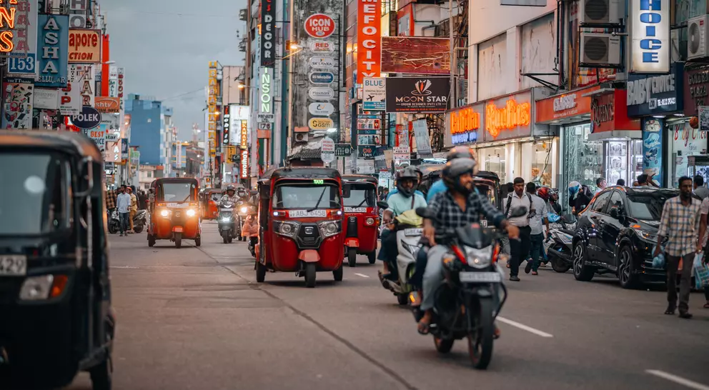 Busy street scene with tuk-tuks, motorcycles, and colorful storefronts illuminated by neon signs.