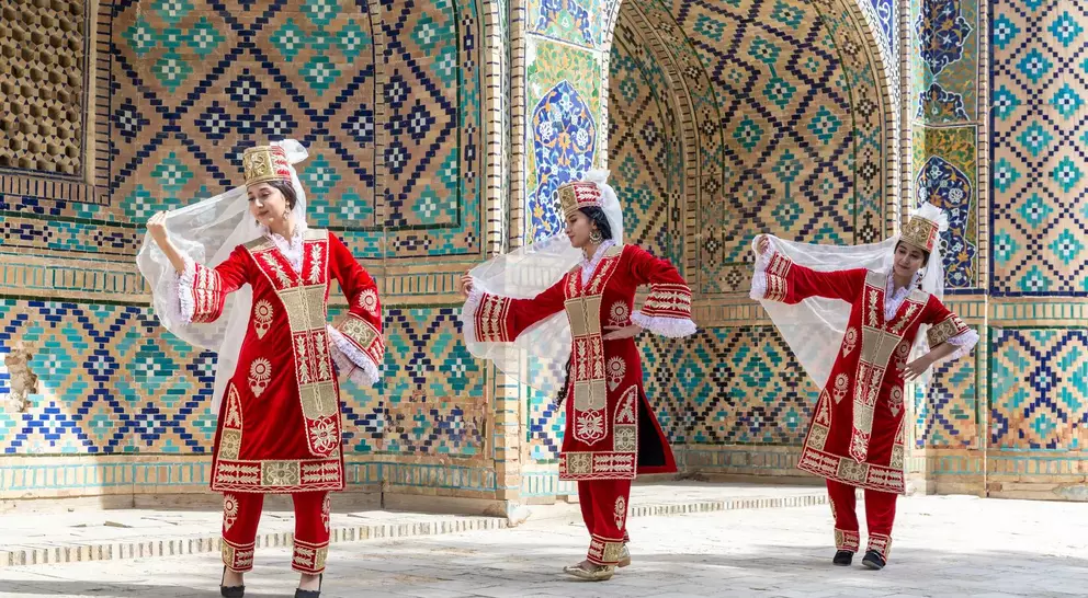 Three dancers in traditional red costumes perform against a colorful, tiled backdrop. They hold flowing white veils.