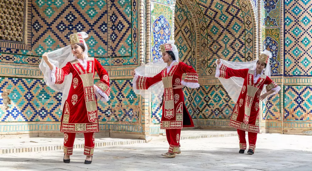 Three dancers in traditional red costumes perform against a colorful, tiled backdrop. They hold flowing white veils.