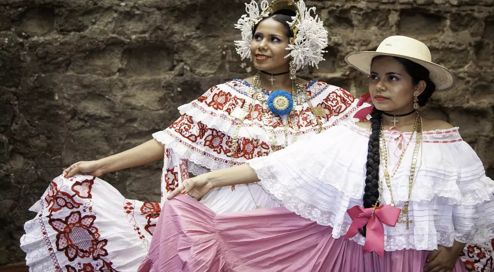 Two women in traditional Mexican dresses pose together, showcasing vibrant colors and cultural accessories against a stone backdrop.