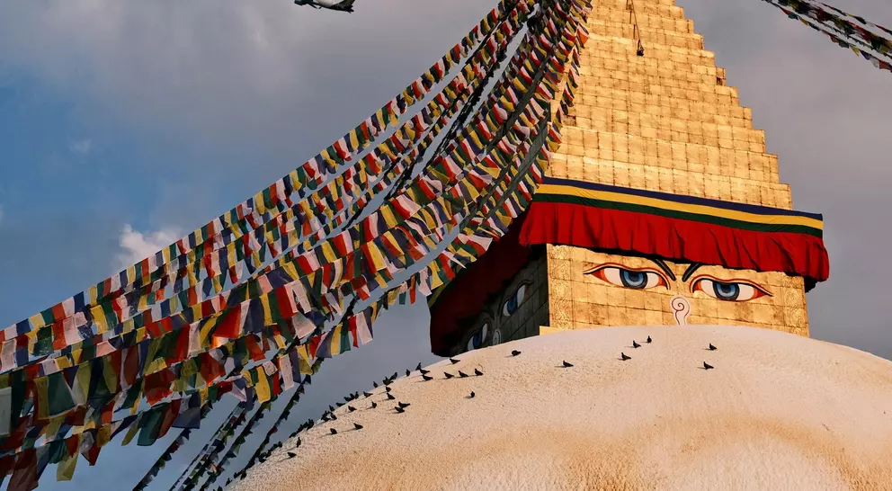 Boudhanath Stupa, adorned with colorful prayer flags, stands under a cloudy sky, with an airplane flying overhead.