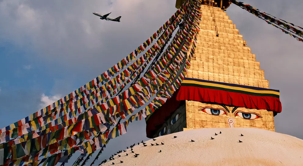 Boudhanath Stupa, adorned with colorful prayer flags, stands under a cloudy sky, with an airplane flying overhead.