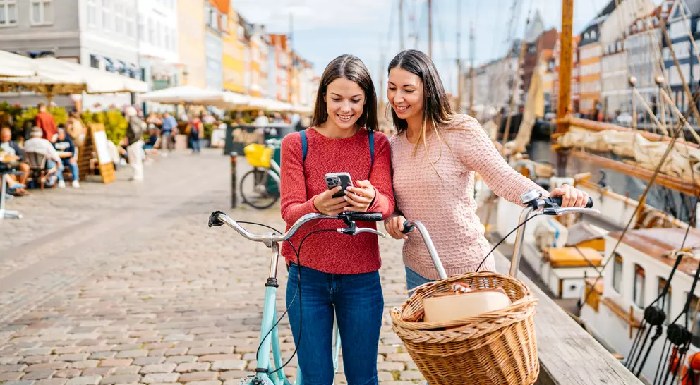 Two women smile while looking at a phone, with a bicycle and scenic harbor in the background.