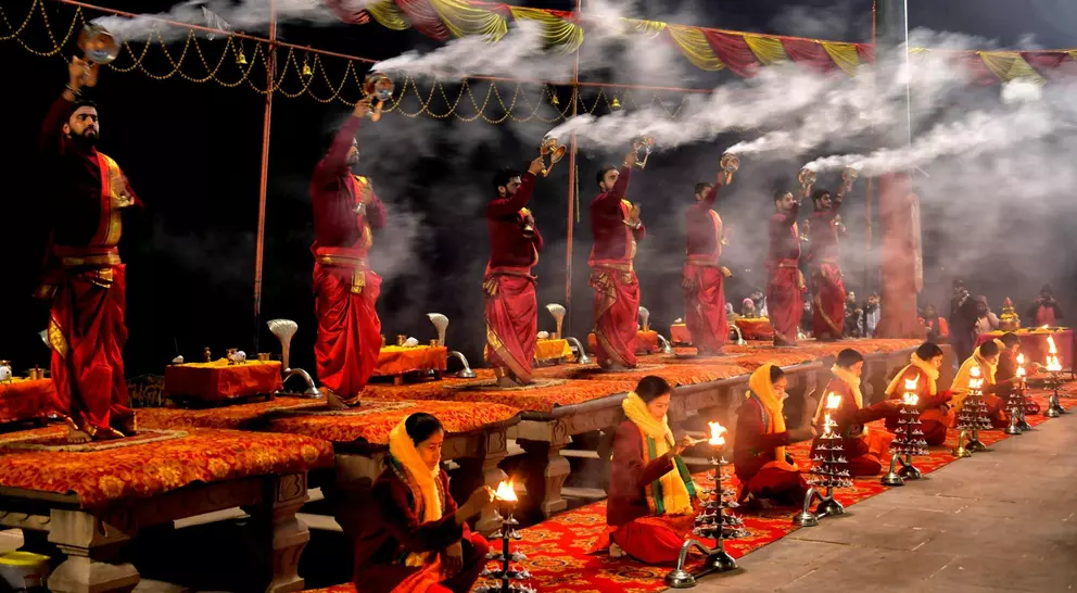 A group of individuals in traditional attire perform a ritual with lamps and incense, surrounded by smoke and bright decorations.