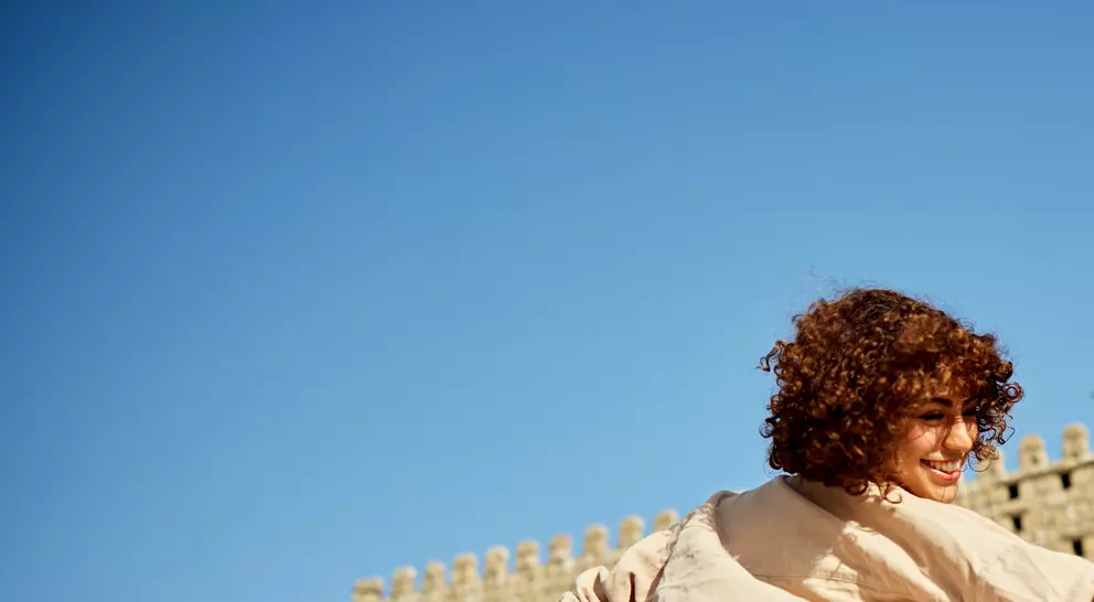 A person with curly hair joyfully spins in front of a large stone archway against a clear blue sky.