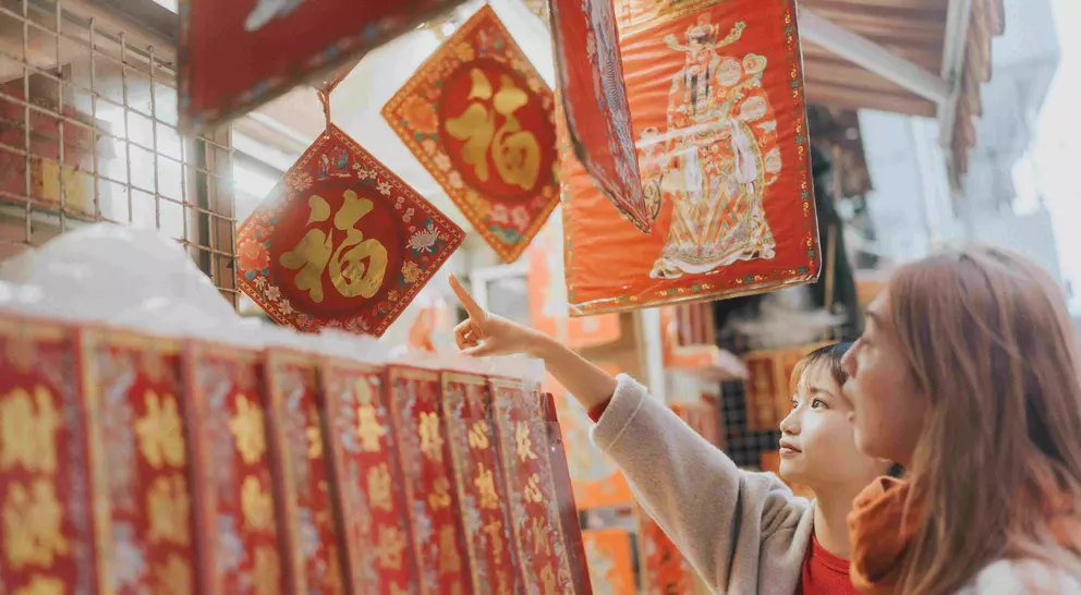 Two women admire decorative red and gold items hanging at a market, pointing at one while browsing the scene.