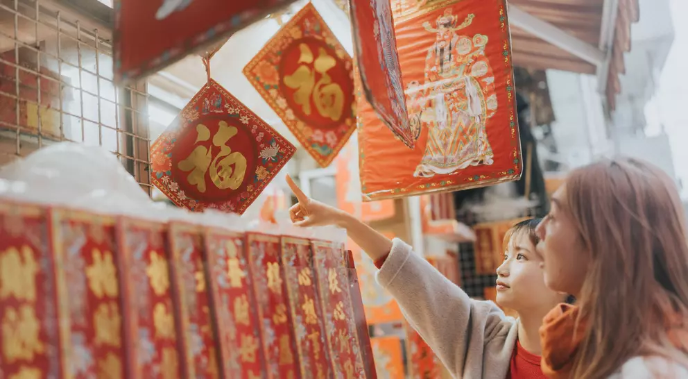 Two women admire decorative red and gold items hanging at a market, pointing at one while browsing the scene.