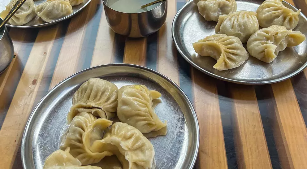 Three metal plates hold steamed dumplings, with a bowl of sauce on a wooden table.