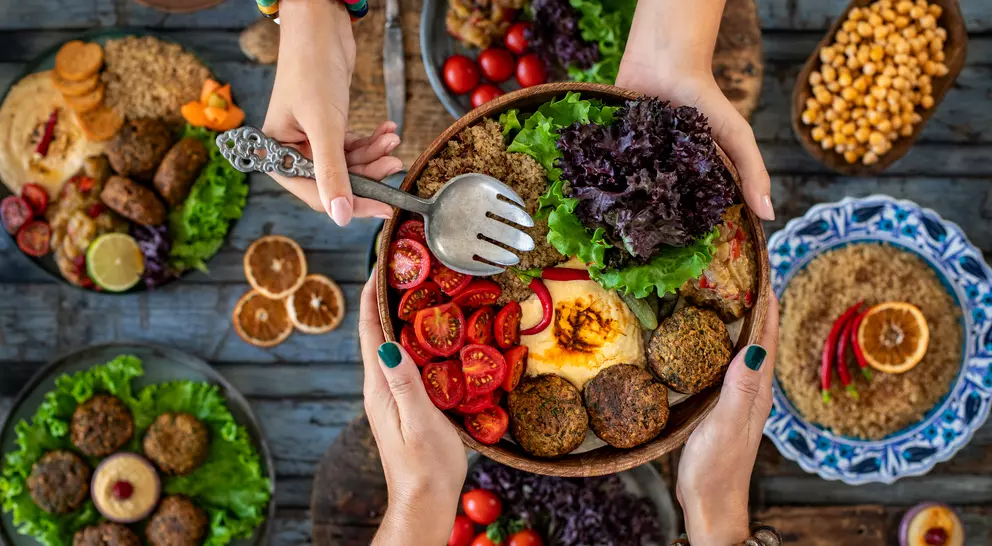 A colorful spread of salads, falafel, and vegetables, with hands reaching for a bowl on a rustic wooden table.