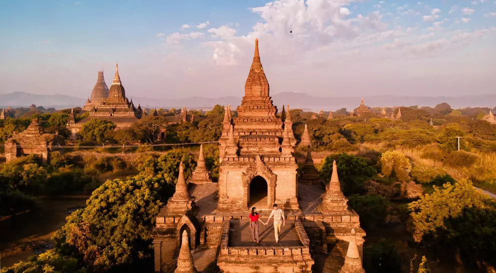 Aerial view of ancient temples in Bagan, Myanmar, with two people exploring among greenery under a blue sky.