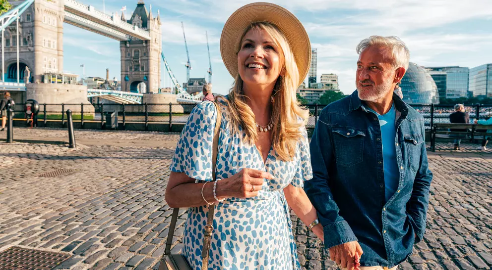 A smiling couple strolls hand in hand near the Tower Bridge in London on a sunny day.
