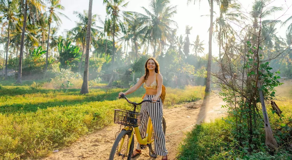 A woman rides a yellow bicycle on a dirt path surrounded by greenery and palm trees, with sunlight filtering through.