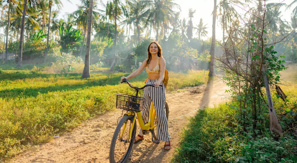 A woman rides a yellow bicycle on a dirt path surrounded by greenery and palm trees, with sunlight filtering through.