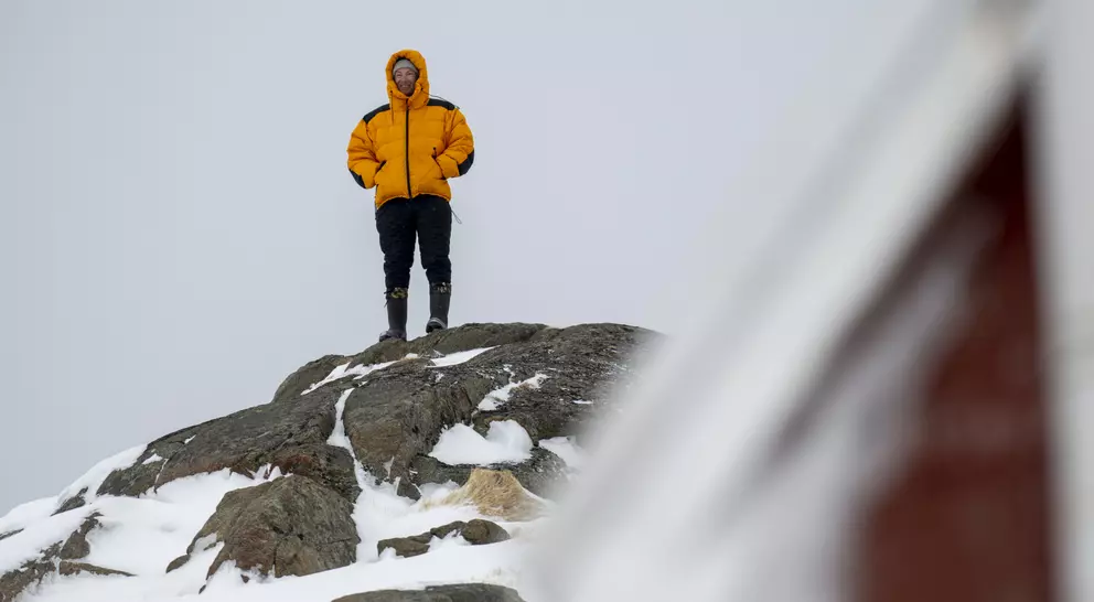 A person in a bright yellow jacket stands on a snowy rock, overlooking a winter landscape with a blurred structure nearby.