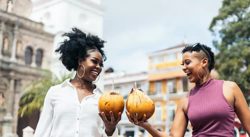 Two women smile and hold coconuts in a vibrant outdoor setting with historic buildings and greenery in the background.