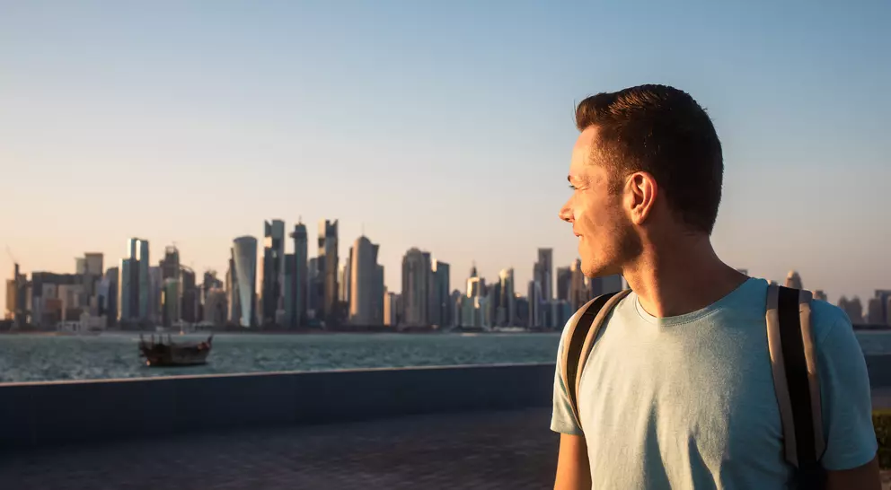 A man gazes at a city skyline during sunset, with water and a boat in the foreground.