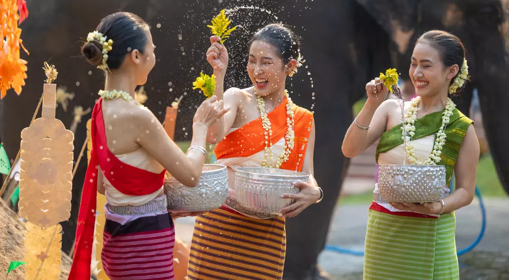 Three women in colorful traditional outfits joyfully splash water while celebrating with an elephant in the background.