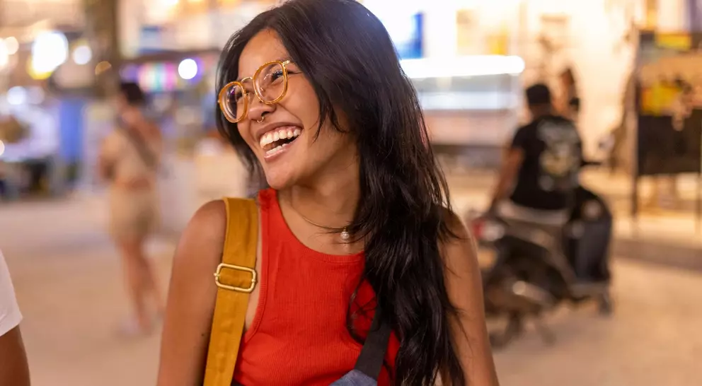 Smiling woman with long black hair, wearing glasses and a red shirt, standing in a bustling outdoor setting.