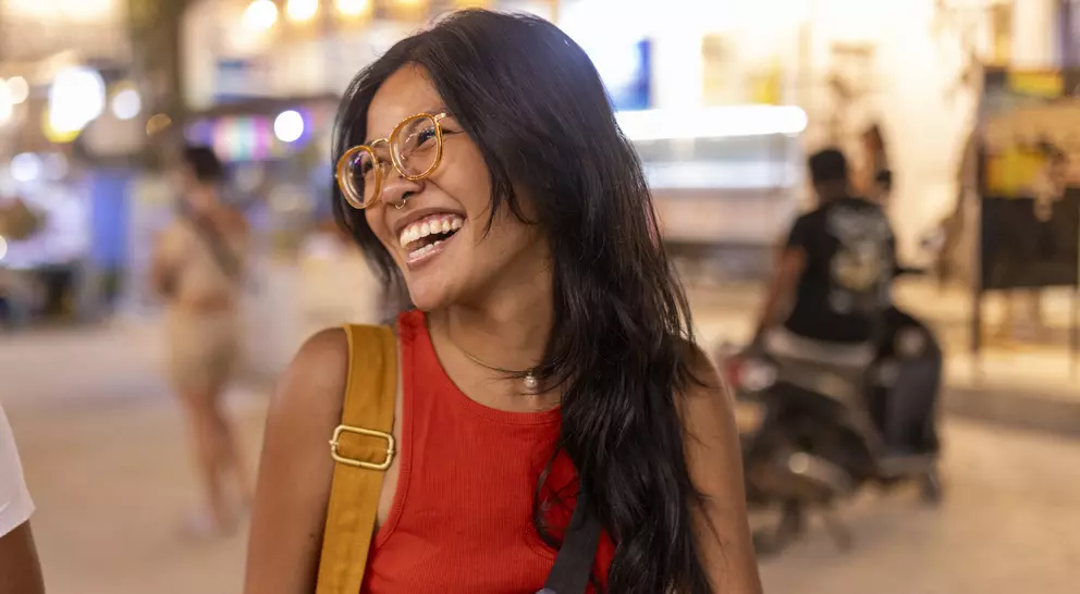 Smiling woman with long black hair, wearing glasses and a red shirt, standing in a bustling outdoor setting.