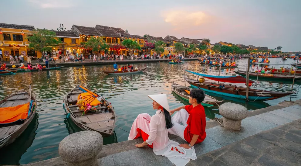 A couple sits by a riverbank in Hoi An, Vietnam, surrounded by boats and lively market scenes at sunset.
