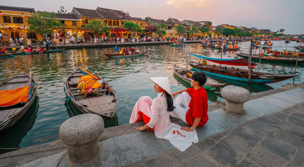 A couple sits by a riverbank in Hoi An, Vietnam, surrounded by boats and lively market scenes at sunset.