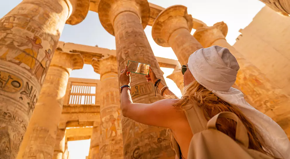 A person taking a photo of ancient columns with hieroglyphs in a sunlit archaeological site.