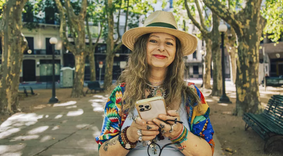 Smiling woman with long, wavy hair, wearing a straw hat and colorful clothing, holding a phone in a sunny park setting.