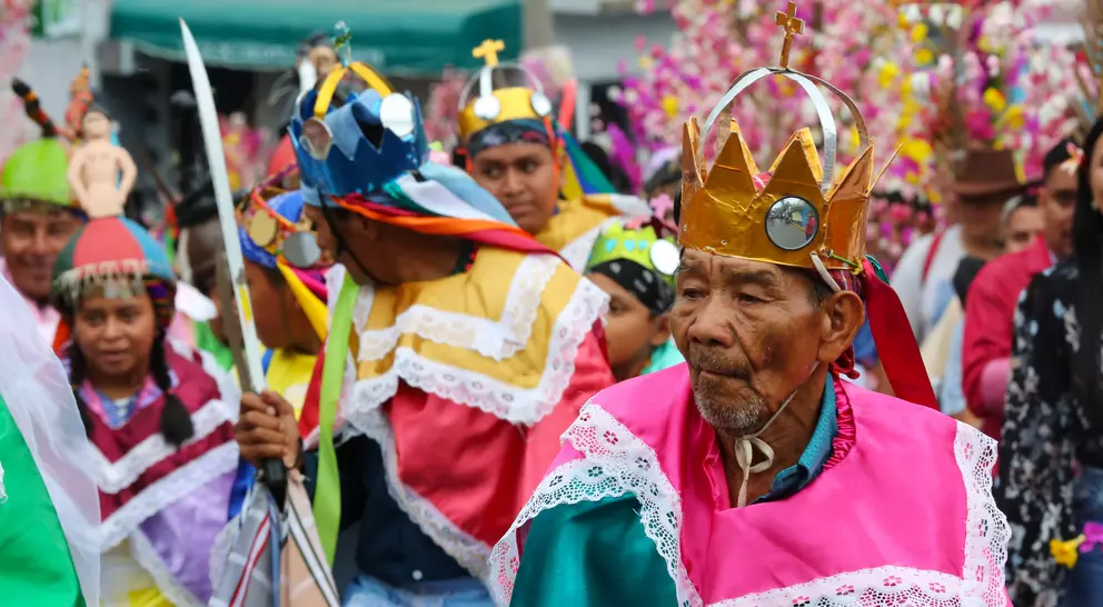 A vibrant parade featuring people in colorful costumes and crowns, celebrating with joyful expressions amidst festive decorations.