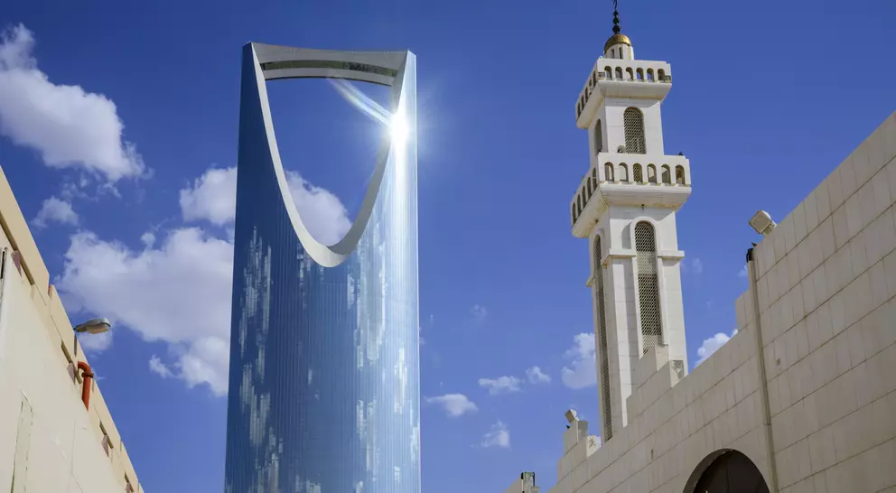 A modern skyscraper with a large opening, next to a traditional mosque tower, against a blue sky with clouds.